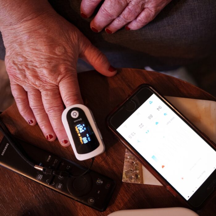 Close up of a hand wearing a fingertip oxygen monitor next to a phone and remote on a table